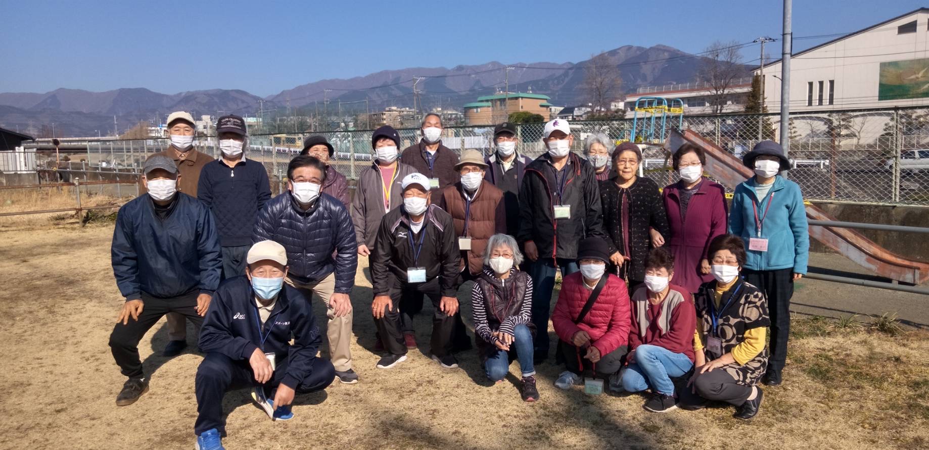 山々が見える公園で老若男女がマスクをして集合して記念撮影している写真