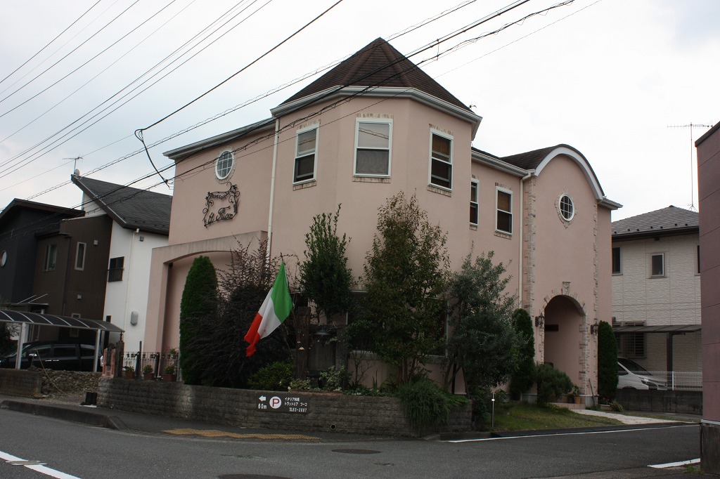 道路の角にピンクベージュの外壁とブラウンの屋根をしたレンガ調のヨーロッパ風建物が建ち、建物の玄関横にはイタリアの国旗が掲げられたお店の外観写真