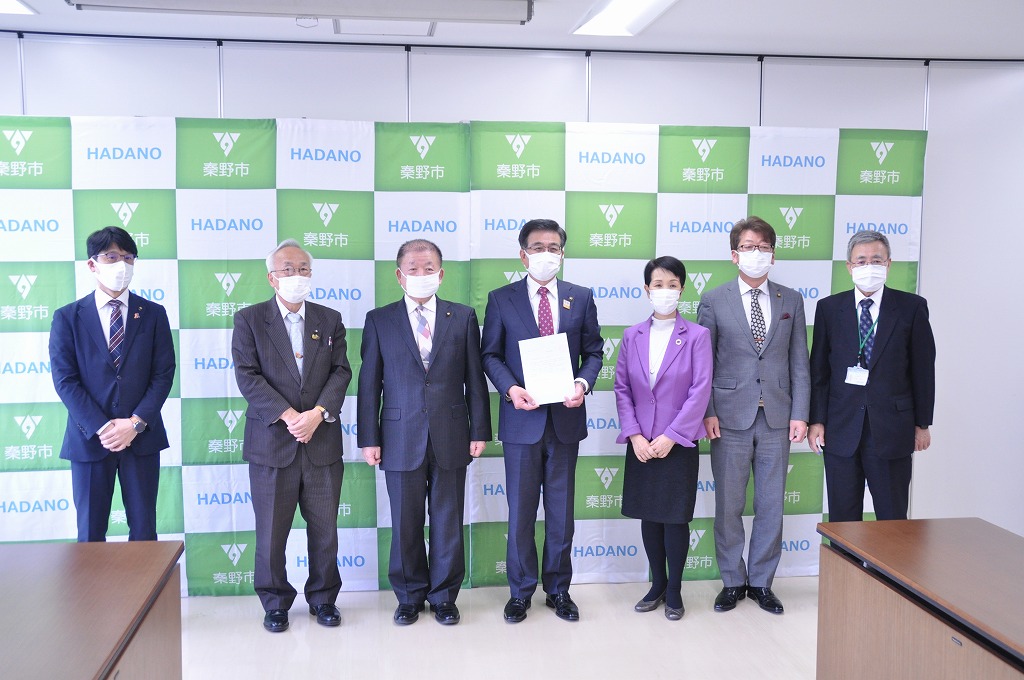 緑と白のチェック柄の旗を背景に、マスクを着用した市長と6人の男女がスーツ姿で要望書を持ち、記念撮影をしている様子の写真