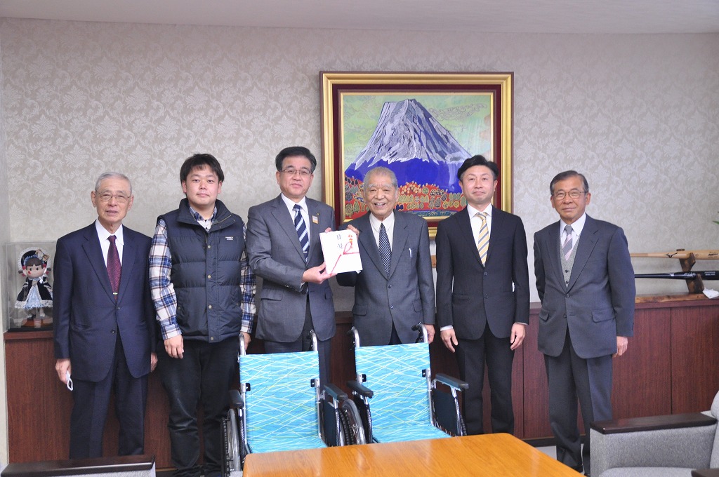 複数のスーツ姿の市長と男性が横一列に並んだ前に、水色の車いすが2台並べられ記念撮影をしている様子の写真