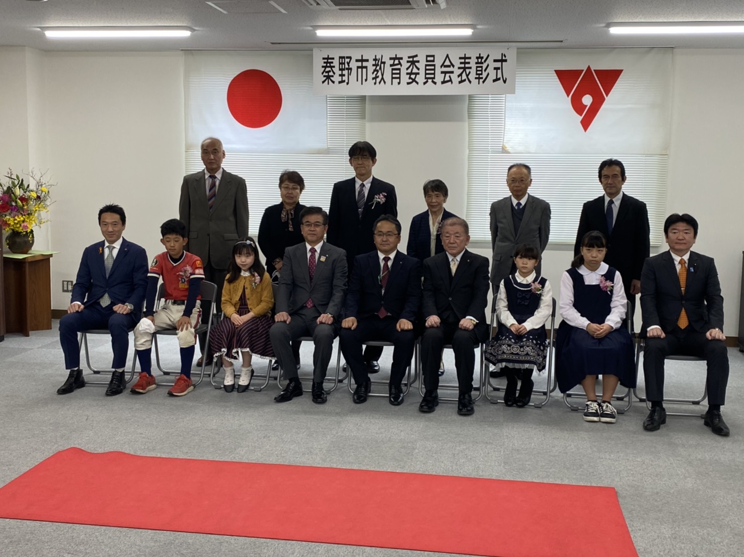 日本国旗や秦野市の旗が掲げられた「秦野市教育委員会表彰式」の横断幕を背景に、受賞者と思われる子どもたちや関係者が記念撮影をしている様子の写真