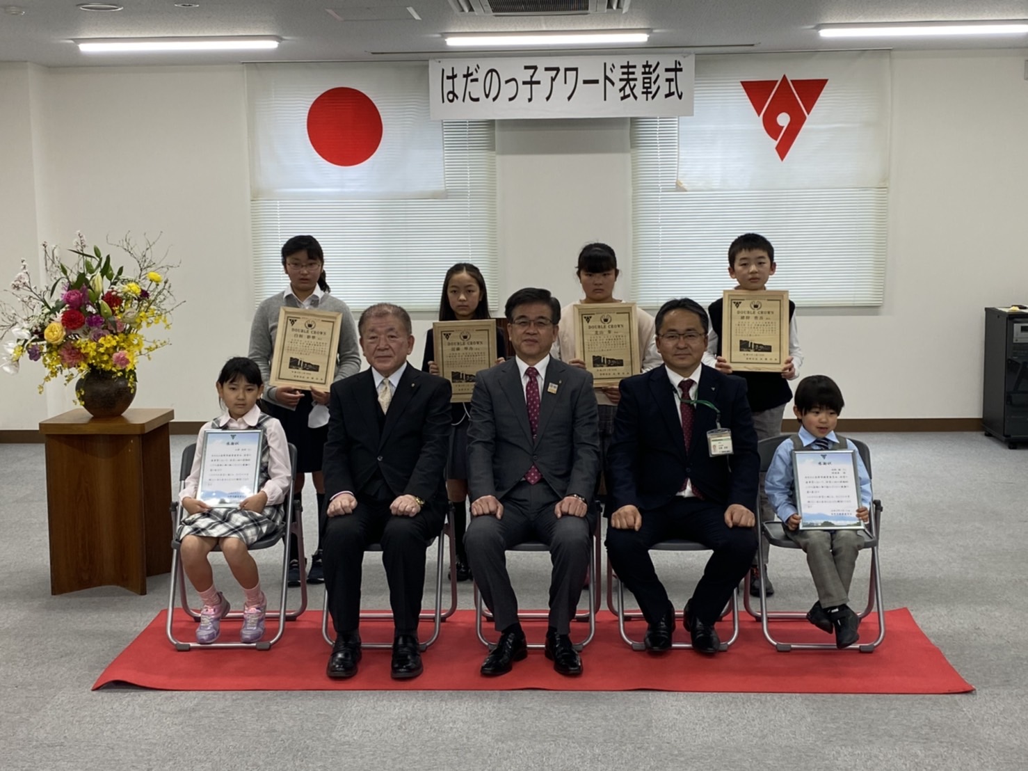 日本国旗や秦野市の旗が掲げられた「はだのっ子アワード表彰式」の会場で、正装した大人3名と表彰状を授与された子ども6名が記念撮影をしている様子の写真