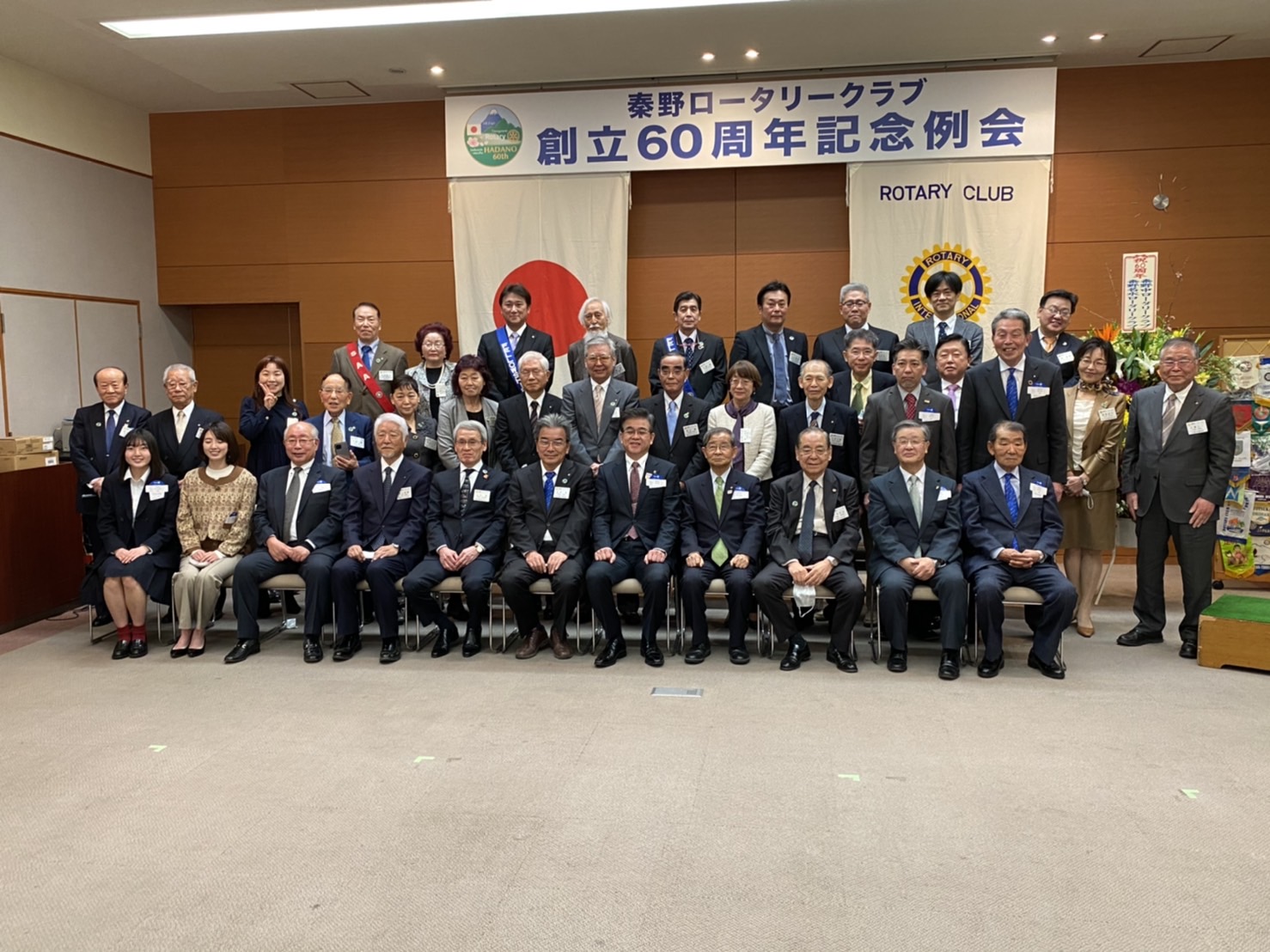 多数の男女がスーツ姿で並び、秦野ロータリークラブ創立60周年記念例会の横断幕を背景に記念撮影をしている様子の写真