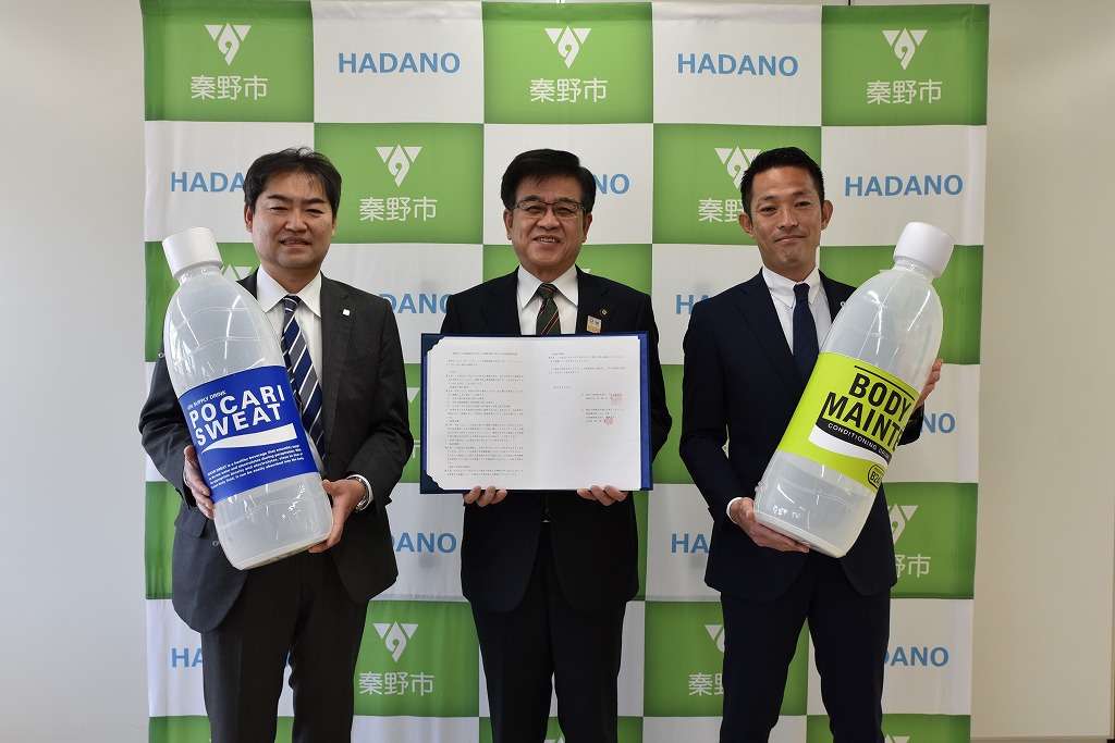 中央の市長は協定書、両側の男性はポカリスエットとボディメンテの巨大なペットボトルを持ち、カメラに向かって立ち記念撮影している写真