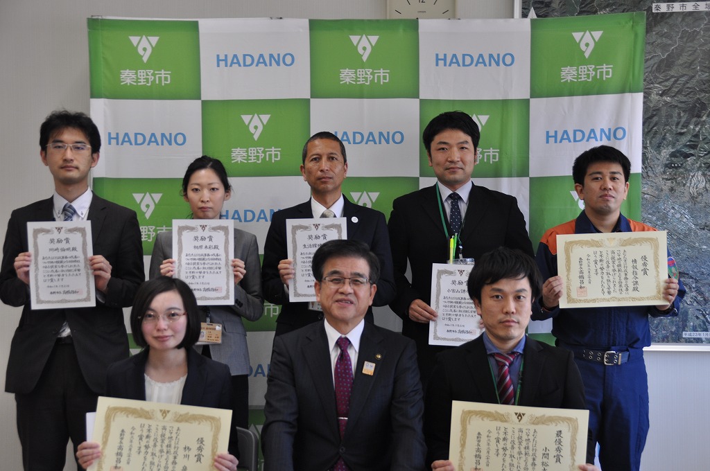 市の旗を背景に、スーツや作業服を着た市長と7人の男女が2列に並び、表彰状を両手で持ち記念撮影をしている写真