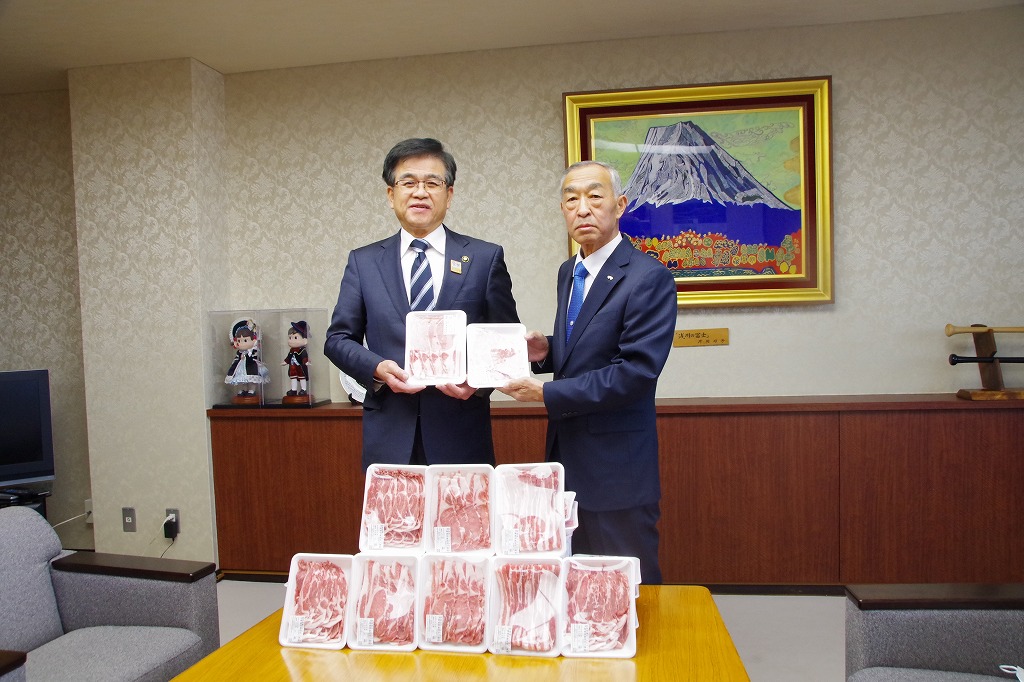 黒いスーツ姿の市長と男性が、テーブルの上に並べられた豚肉のパックを持って記念撮影をしている写真