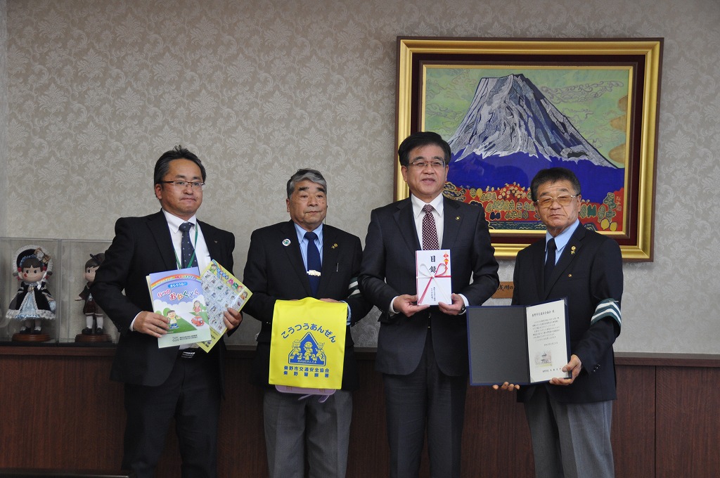 黒いスーツを着た市長と3人の男性が横一列に並び、黄色のランドセレカバーを付けたランドセルや冊紙を持ち記念撮影をしている写真