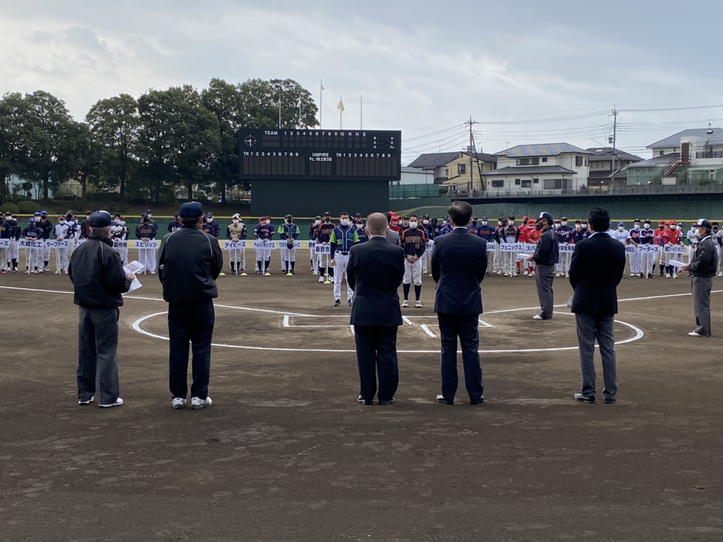 野球場で、多くの野球チームが整列し、背広を着た関係者たちがグラウンドの真ん中で整列している開会式の様子の写真