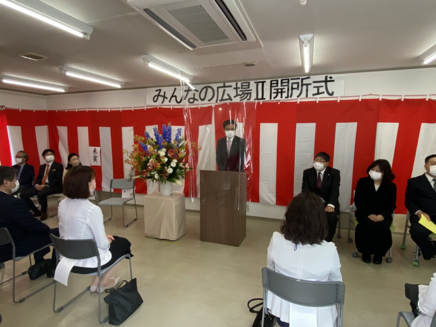 紅白の幕が飾られた会場で、マスクを着用した市長が演台から挨拶をしており、その周囲には複数の来賓や関係者が着席している式典の様子を捉えた写真