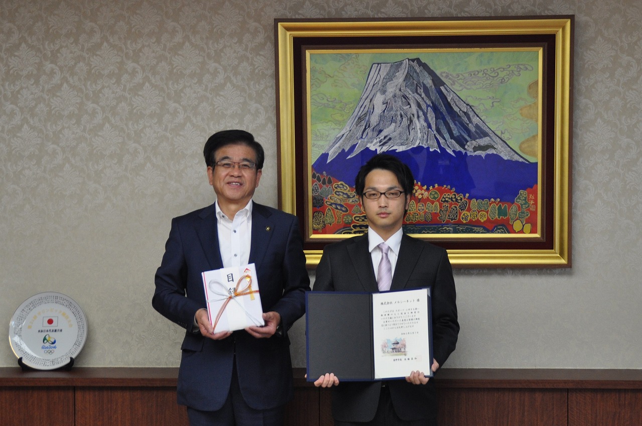 市の応接室でスーツ姿の市長と男性が、目録と感謝状の入ったファイルをそれぞれ持ち、カメラに向かって並んでいる記念写真