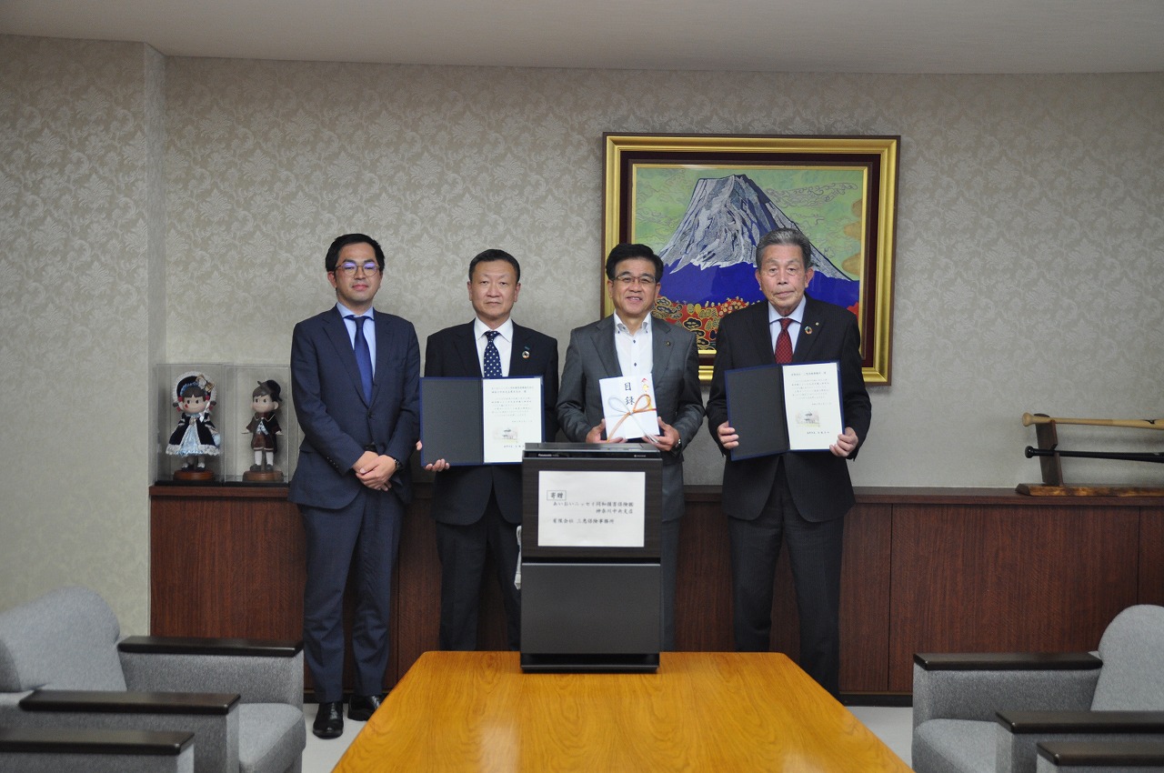 室内の応接室で、スーツ姿の市長と男性3人が、それぞれ賞状や目録などを手に持ち、中央のテーブルには寄附された空気清浄機が置かれ、全員がカメラに向かって並んでいる記念写真