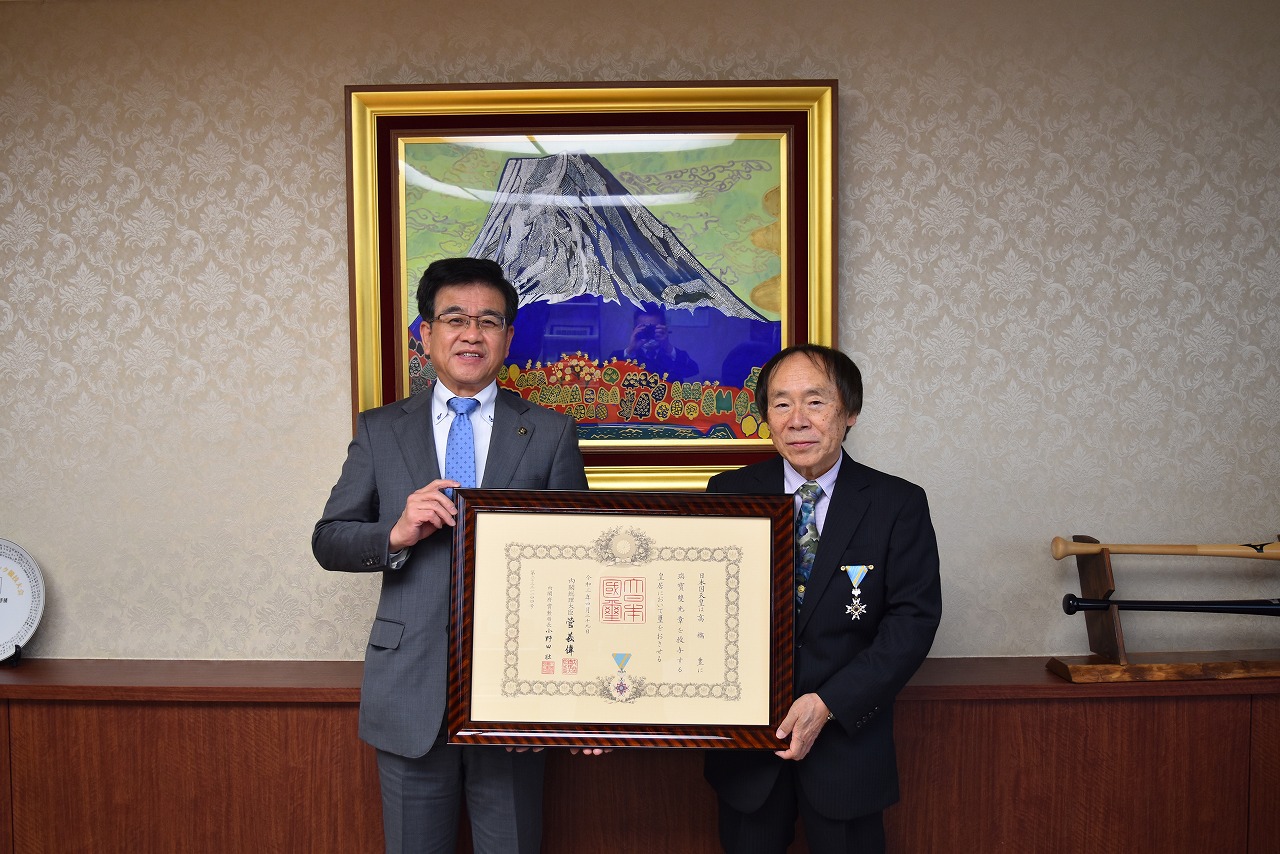 スーツ姿の市長と男性がカメラに向かって並んでおり、勲章と賞状の入った額縁を掲げている写真