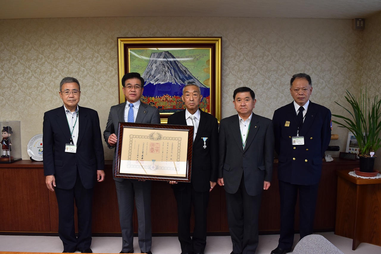 富士山の絵画が飾られた応接室で、スーツ姿の市長と男性4人がカメラに向かって並んでおり、中央の2人が勲章と賞状の入った額縁を掲げている集合写真