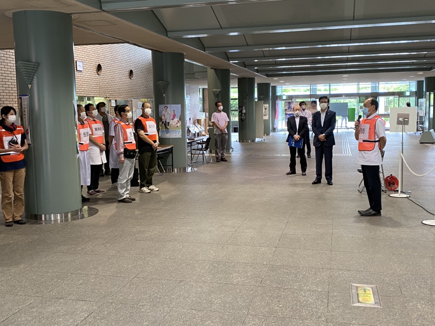 オレンジ色のベストを着用した人々やスーツ姿の男性たちが整列し、男性が全員に話しかけている視察会場での写真