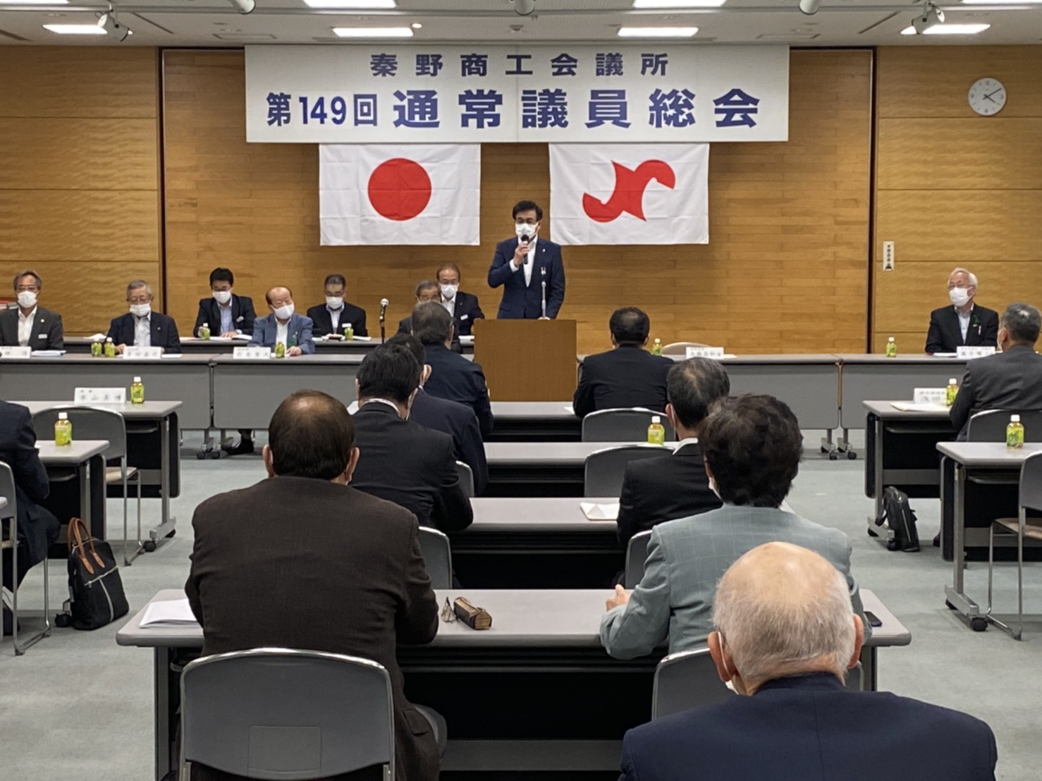 壇上に立つ市長が挨拶しており、多くの参加者が横一列に並んだテーブル席で話を聞いている会場の写真