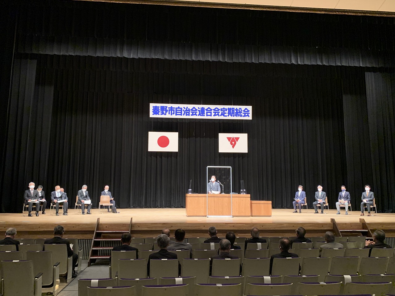 舞台の演台に立つ市長が多くの聴衆を前に挨拶しており、ステージ上には「秦野市自治会連合会定期総会」と書かれた横断幕と日本の国旗が掲げられている総会の写真