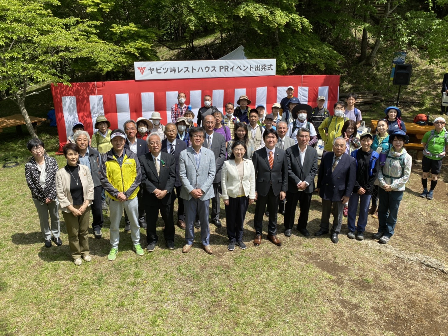 緑豊かな山を背景に、多くの人々が「ヤビツ峠レストハウスPRイベント出発式」と書かれた赤い横断幕の前でカメラに笑顔で集合している屋外での写真