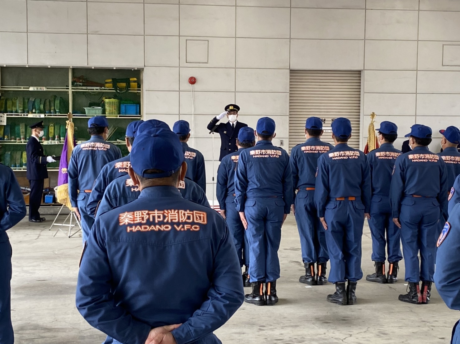屋内の広場で、「秦野消防団」と書かれた紺色の制服を着た人々が整列し、その前で制服姿の男性が敬礼している表彰式での写真