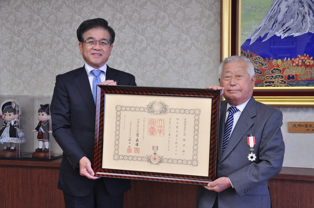 室内で、スーツを着た市長と男性が額縁を一緒に両手で持ち、記念撮影している写真