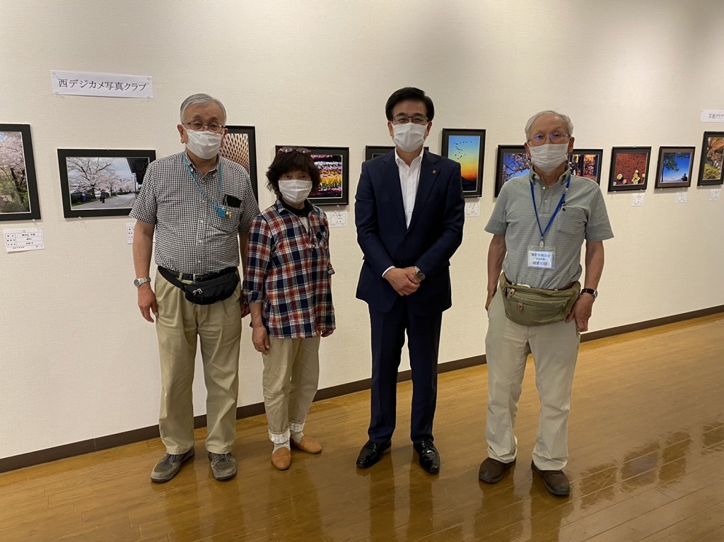 額に入れられた複数の写真が壁に展示されている会場で、マスクを着用した市長と男女3人が横一列に並び記念撮影している写真