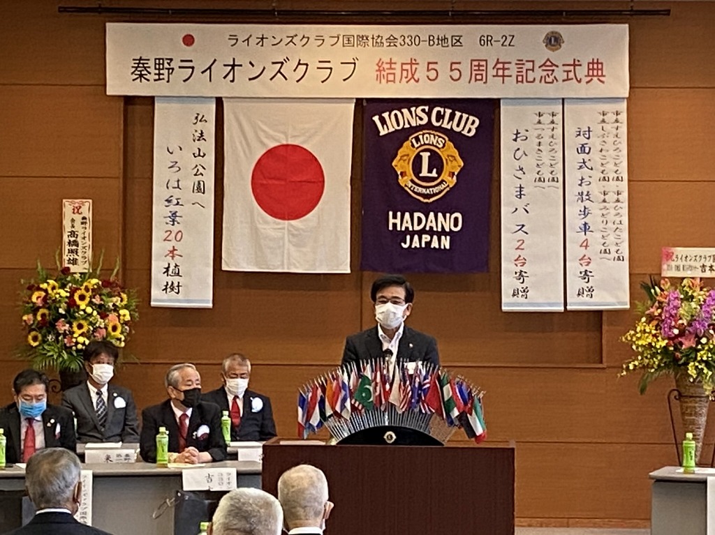 横断幕や国旗、団旗、垂れ幕が掲げられたステージ上で複数の国旗が飾られている演台にマスクを着用した市長がスピーチをしている写真