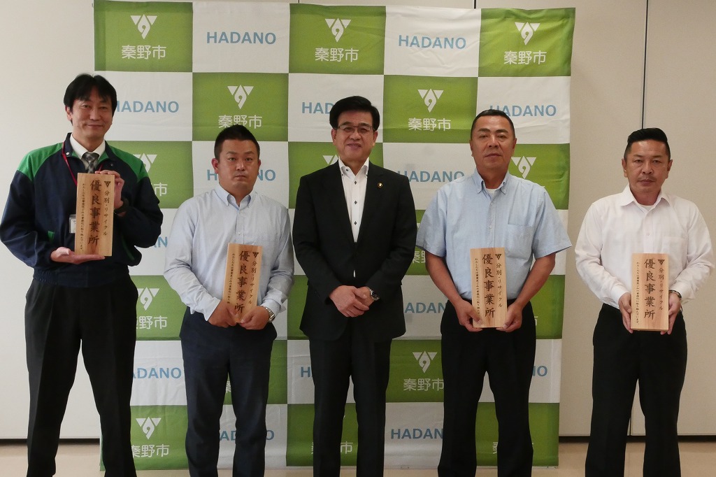 秦野市のバックボードの前でスーツを着た市長と作業着風の男性を含めた5人の男性が並んでおり、それぞれが「優良事業所」と書かれた木製の表彰盾を持っている集合写真