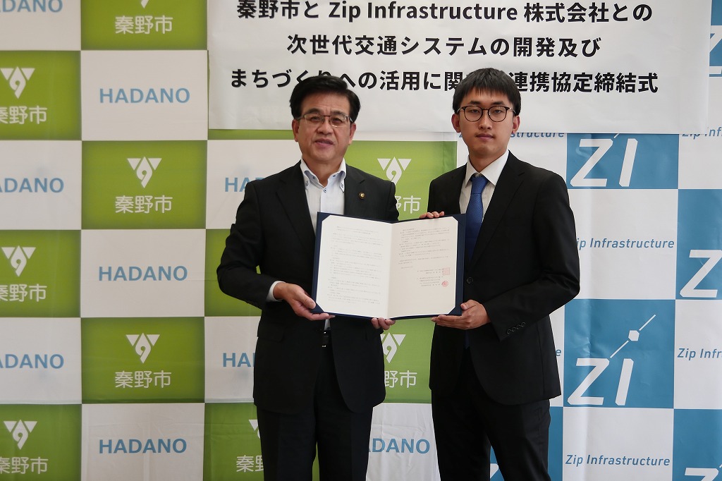 秦野市とZip Infrastructure株式会社のバックボードの前でスーツ姿の市長と男性が協定書も一緒に持ち記念撮影している写真