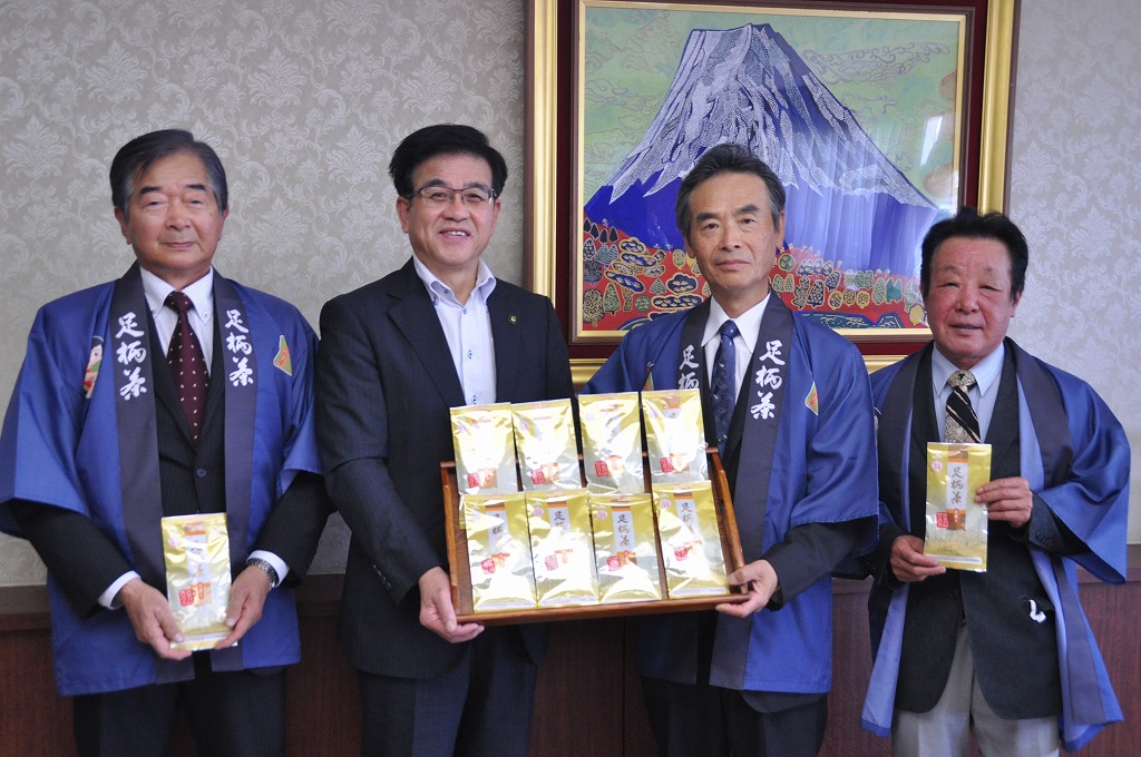 青い法被を着用した3人の男性とスーツ姿の市長が、お茶が入った箱を持ち記念撮影をしている写真