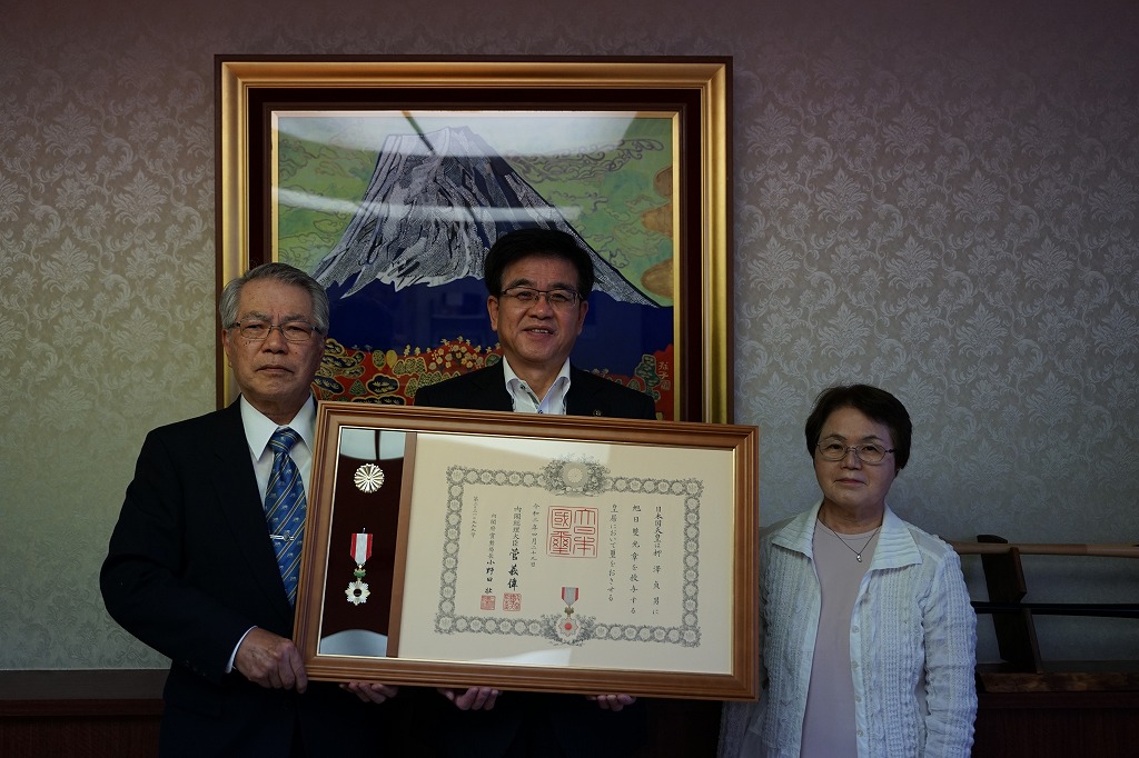 富士山の絵画が飾られた応接室で、スーツ姿の男性と女性に挟まれた市長が、勲章と感謝状の入った大きな額縁を持って微笑んでいる写真