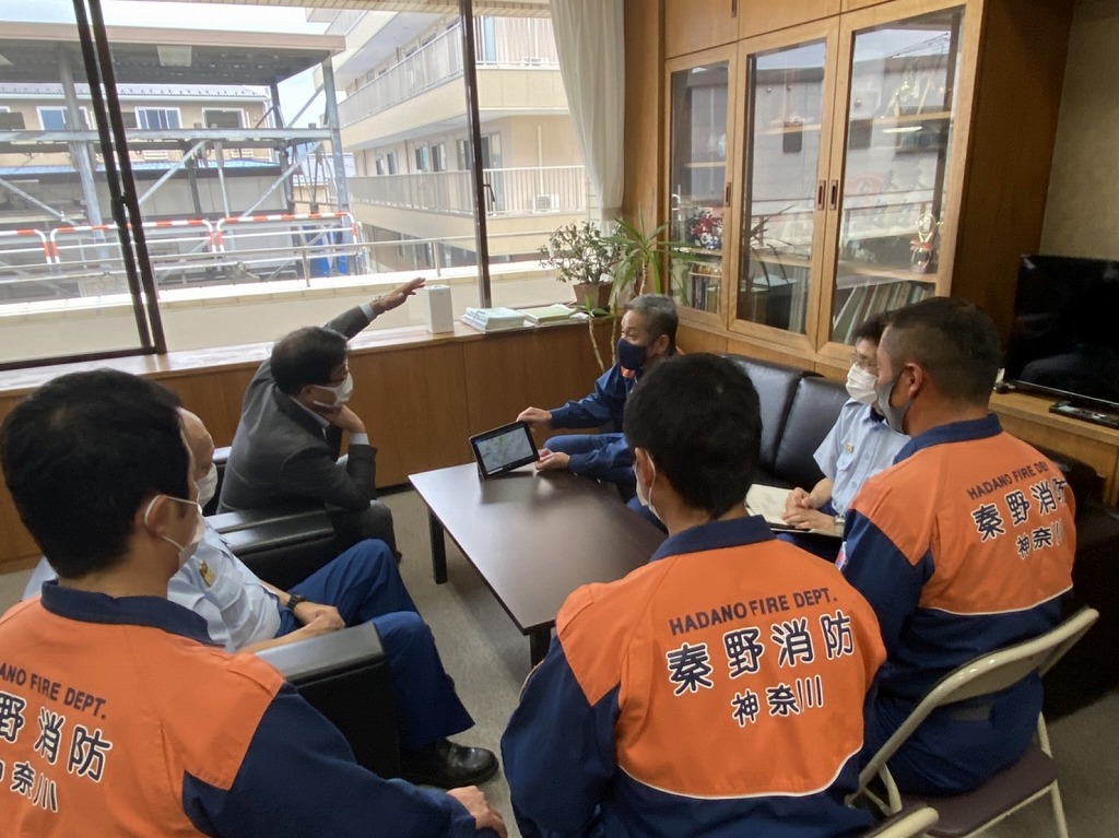 オレンジ色の作業服を着た消防士とスーツ姿の男性がテーブルを囲んでタブレット端末の画面を見ながら活動報告をしている写真