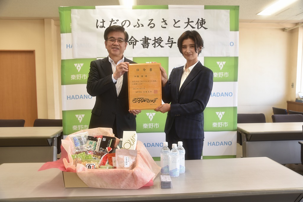 スーツ姿の市長と女性が「はだのふるさと大使」と書かれた木製の任命書をカメラに向かって一緒に掲げており、手前には地元の特産品が並んでいる写真