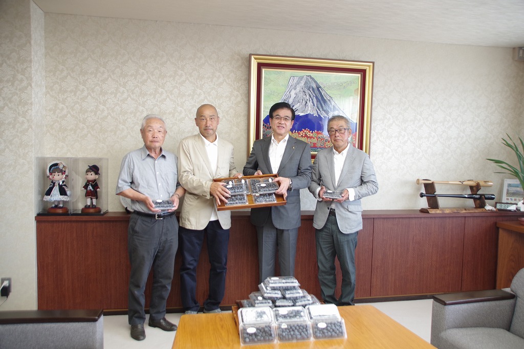 スーツ姿の市と3人の男性が、積み重ねられたブルーベリーのパックや感謝状をカメラに向かって掲げている室内の写真