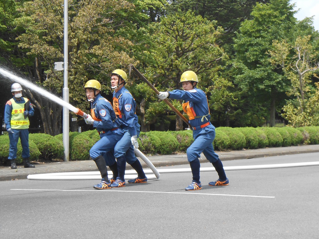 黄色いヘルメットと青い訓練服を着た消防士たちが、放水や器具の操作をしながら道路上で消防訓練を行っている写真