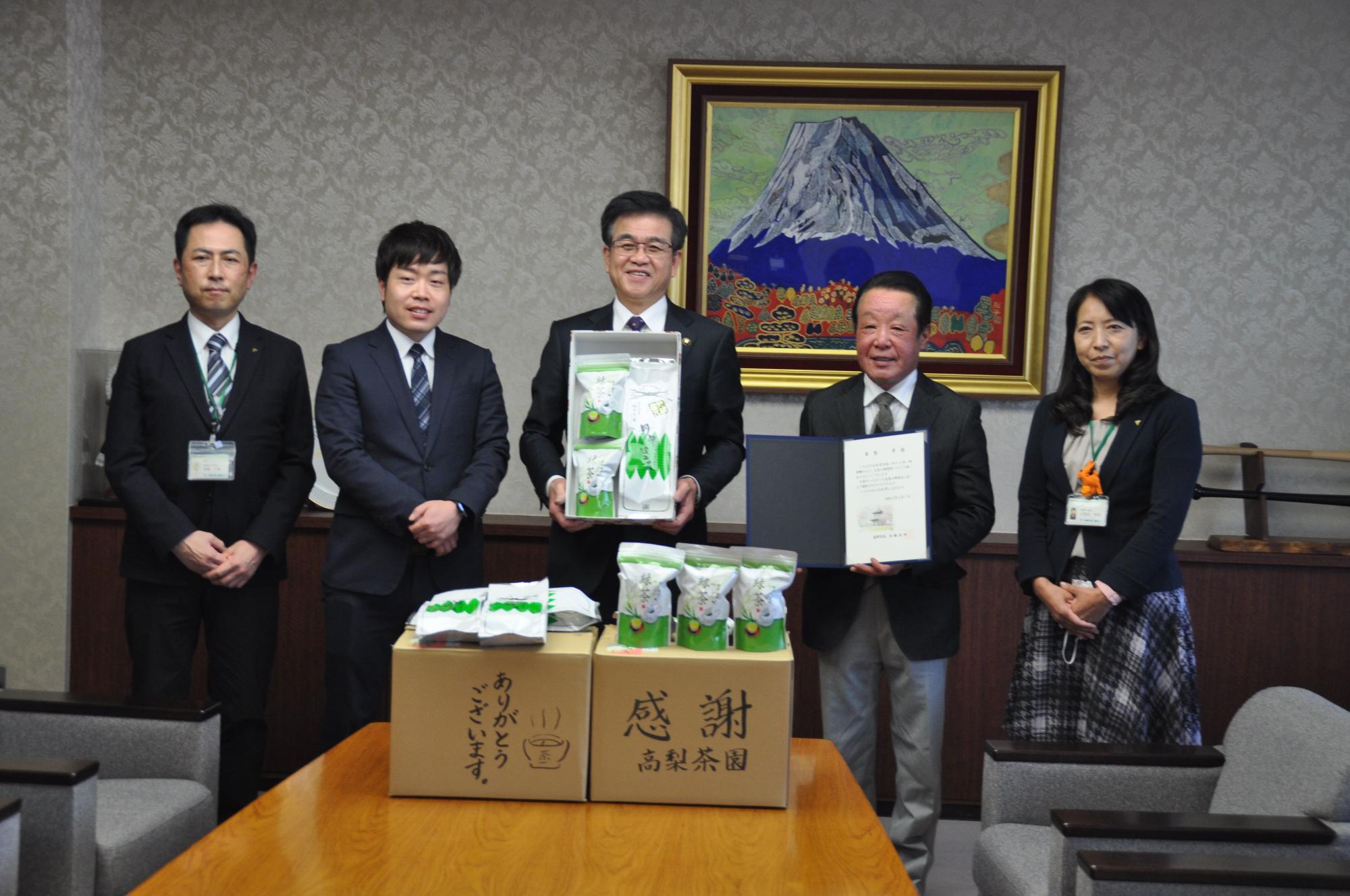 富士山の絵画が飾られた部屋で、テーブルの茶葉の箱を前に、市長が茶葉の箱を持ち、その両脇に感謝状を持ち男女4人が並んで写っている贈呈式の写真