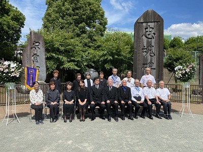 たくさんの人が椅子に座ったり立ったりして並び、その奥には「忠魂碑」と「永世之碑」という2つの大きな石碑が立っている追悼式の写真