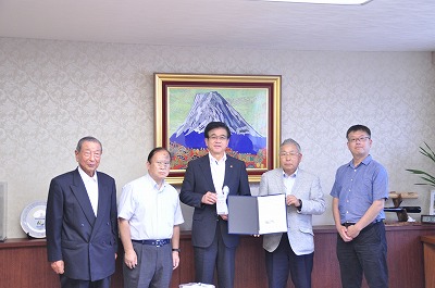 富士山の絵画が飾られた部屋で、中央の市長は目録、右側の男性は書類を持って並んでいるスーツ姿やワイシャツ姿の5人の男性の写真