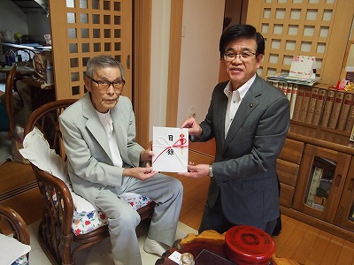 和風の部屋で、籐椅子に座った高齢の男性とスーツ姿の市長が、目録と書かれた熨斗袋を一緒に持っている写真