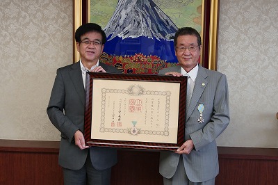 富士山の絵画が飾られた部屋で、スーツ姿の市長と男性が、中央で勲章のついた大きな額縁を並んで持っている写真