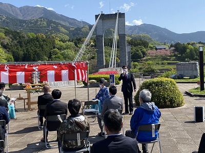 山の斜面に立つ巨大な吊り橋を背景に、紅白幕の祭壇と参加者たちが写っており、スーツ姿の市長がマイクで挨拶をしている写真