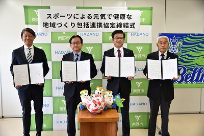 緑と白のロゴのバックパネルを背景に、「スポーツによる元気で健康な地域づくり包括連携協定締結式」の看板の下で、市長と3人の男性が協定書を持ち、中央のテーブルにはサッカーボールと複数のマスコット人形が置かれている写真