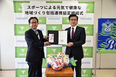 「スポーツによる元気で健康な地域づくり包括連携協定締結式」の看板の前で、スーツ姿の市長と男性で協定書と熨斗袋を持ち、中央のテーブルにはマスコットとサッカーボールが置かれている写真