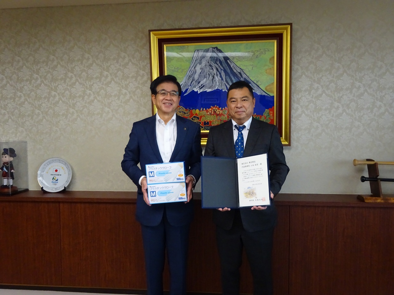 富士山の絵画を背景にした部屋で、スーツ姿の市長はゴム手袋の箱を、もう1人の男性は書類を持って並んでいる写真