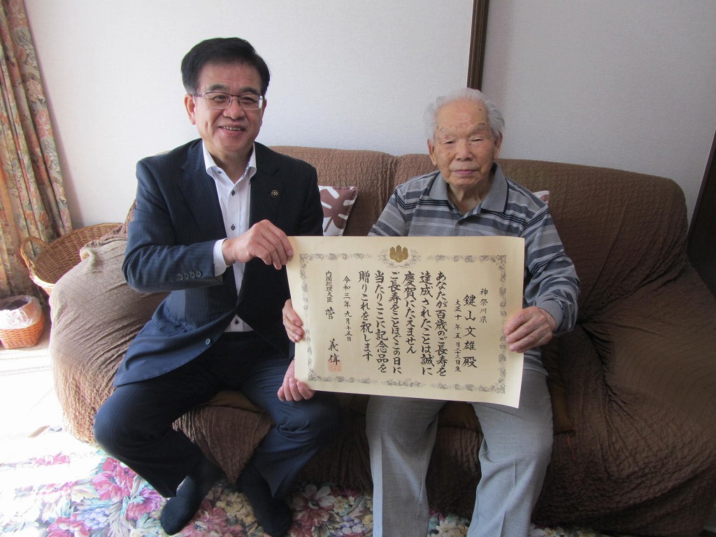 長寿を祝う賞状を中央で持ち、市長と男性がソファに並んで座り、笑顔でカメラを見つめている写真