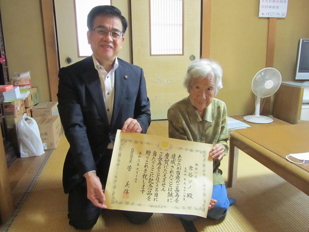 座敷でスーツ姿の市長が女性と並んで正座しており、二人で長寿を祝う賞状を掲げている写真