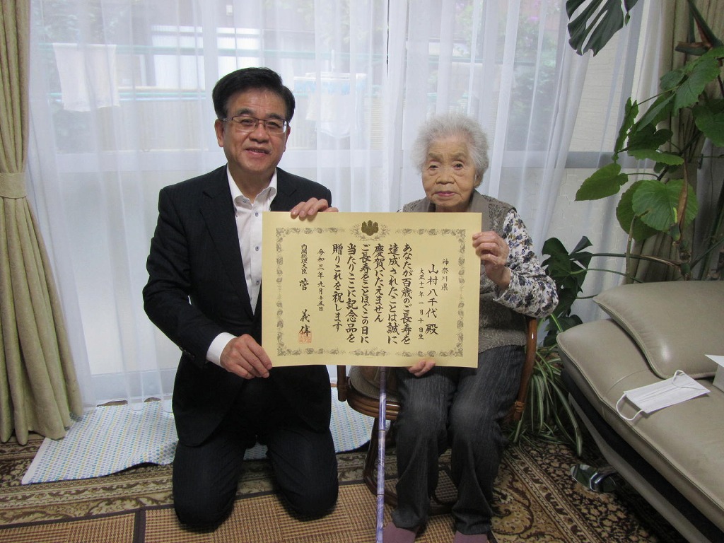 和室で、スーツ姿の市長が正座し、椅子に座った女性と共に、長寿を祝う賞状を掲げてカメラに笑顔を向けている写真