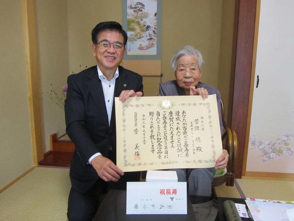 和室で高齢女性とスーツ姿の市長が、長寿を祝う賞状と記念品を囲んで笑顔でカメラを見つめている記念写真