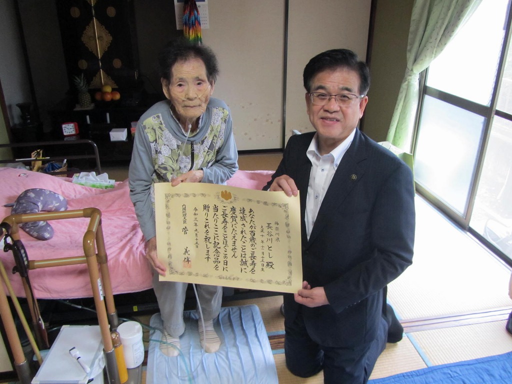 和室でスーツ姿の市長が膝立ちし、高齢の女性が介護ベッドに座って長寿を祝う賞状を掲げている記念写真