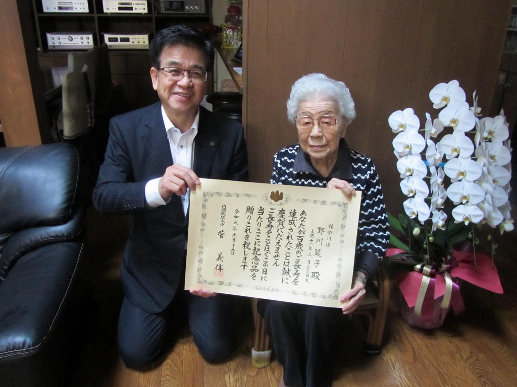 フローリングの部屋で、スーツ姿の市長が正座し、椅子に座った高齢の女性と共に、白い胡蝶蘭の鉢植えを囲んで長寿祝いの賞状を掲げている写真