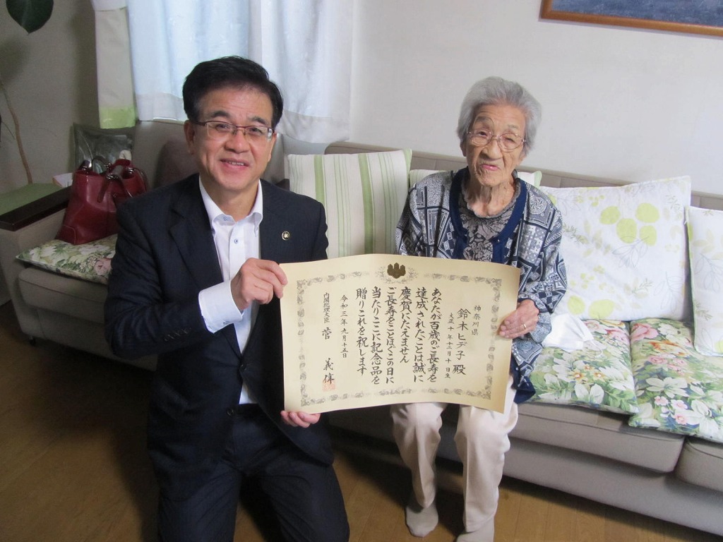 スーツ姿の市長が正座し、ソファに座った高齢の女性と共に、長寿祝いの賞状を笑顔で掲げている写真