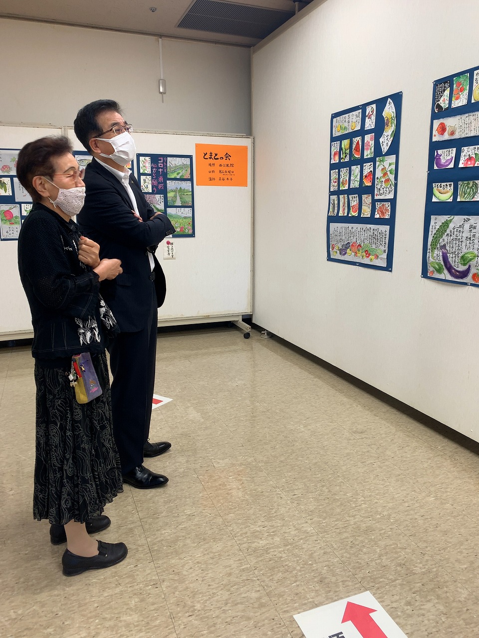 マスクを着用した市長と女性が、壁に展示されている野菜や果物の絵画を熱心に鑑賞している写真